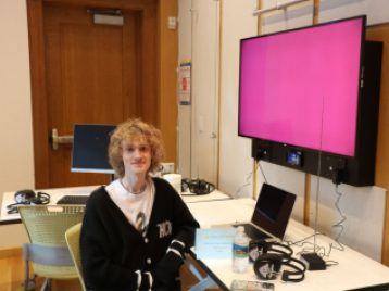 Jameson Van Dokkumburg sits beside a computer, sets of headphones, and a monitor.
